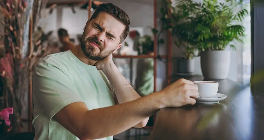 A man in a green shirt is grimacing and holding his neck in pain during his lunch break and requires workers' comp medical evaluation for workplace injury in North Carolina.