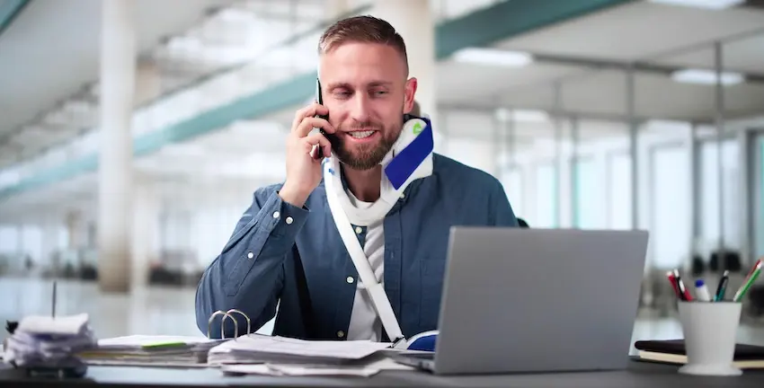 An injured worker in a neck brace and arm sling is on the phone with a laptop and is filing a workers' comp claim from an office in North Carolina.
