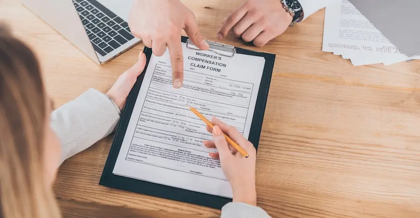 Two people are reviewing and completing a workers' comp claim form on a clipboard at a wooden desk with a laptop and documents at 1 Charlotte Injury Lawyers in North Carolina.