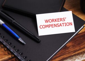 A white card with red workers' compensation text on a black notebook beside pens representing workplace injury benefits claim in North Carolina