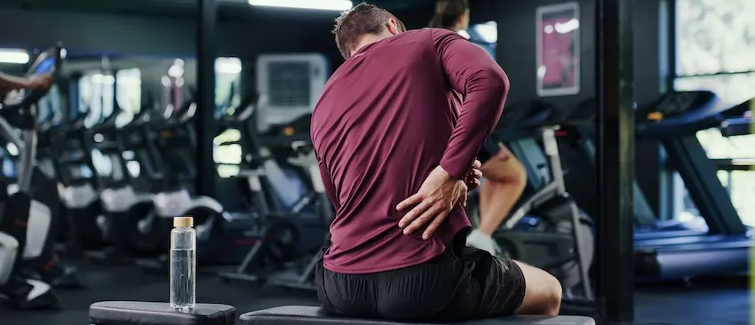 A man in a maroon long-sleeve shirt sits on a gym bench grabbing his lower back in pain while surrounded by cardio equipment depicting the type of premises liability injury that a personal injury lawyer from 1 Charlotte Injury Lawyers can help victims pursue compensation for.