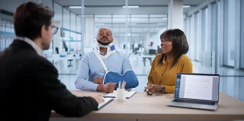 A man wearing a neck brace and arm sling sits beside a woman in a yellow blouse across from an attorney taking notes at a modern office conference table capturing the initial consultation process when an injured client and their spouse meet with a personal injury lawyer from 1 Charlotte Injury Lawyers to discuss their case in North Carolina.