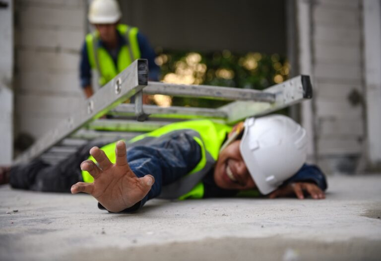 A construction worker in a yellow safety vest and white hard hat lies face-down on a concrete floor with his arm outstretched and an aluminum ladder collapsed on top of him while a coworker in the background rushes toward him capturing the dangerous aftermath of a ladder fall construction accident on an active job site in North Carolina.