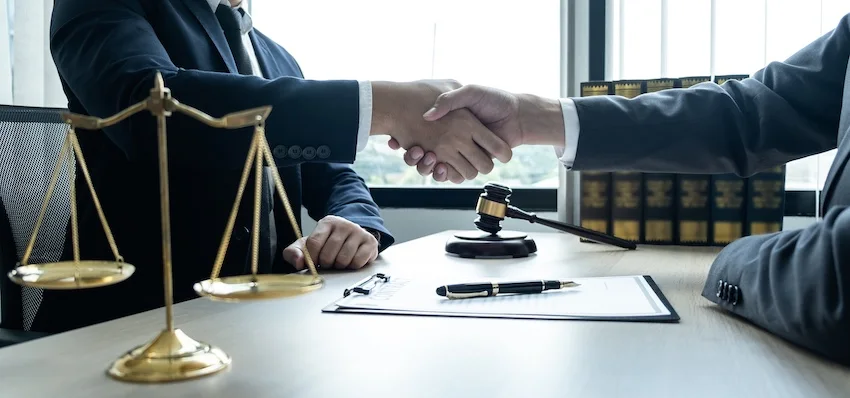 An attorney from 1 Charlotte Injury Lawyers in dark suits shakes hands with a client across a desk beside a gold scale of justice- a gavel- and legal documents- representing the moment an injured cyclist formally retains a bicycle accident lawyer to pursue compensation after a crash.