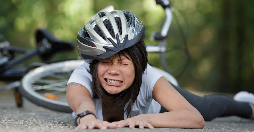A female cyclist wearing a helmet lies on the ground in pain with her bicycle tipped over behind her- depicting the type of serious fall or collision that leads injured riders to contact a bicycle accident lawyer from 1 Charlotte Injury Lawyers for help recovering damages.