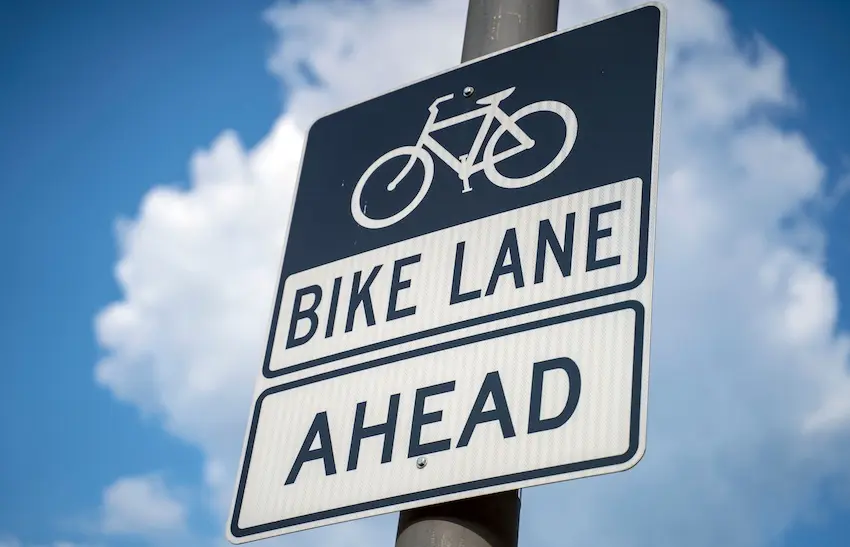 A blue and white road sign mounted on a pole against a partly cloudy sky reads "Bike Lane Ahead" with a bicycle icon at the top- representing the designated cycling infrastructure whose violation by drivers often forms the basis of a bicycle accident lawyer's negligence case in Charlotte.