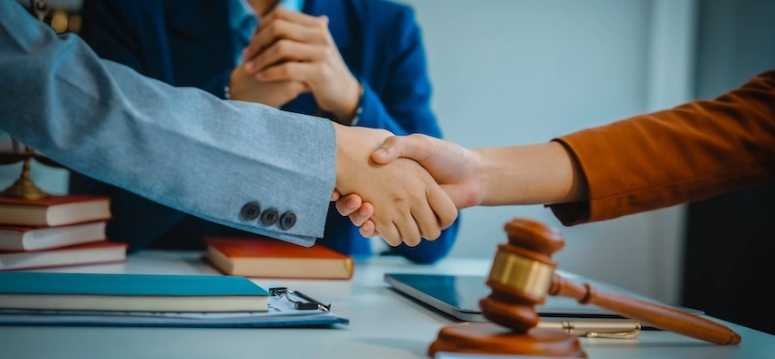 A client and attorney from 1 Charlotte Injury Lawyers shake hands across a desk with a gavel- clipboard- and law books in view while a third person sits with folded hands in the background- representing the moment an injured rider formally retains a motorcycle accident lawyer to handle their injury claim.